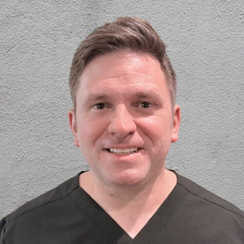 Smiling man in black scrubs standing against a textured gray wall.